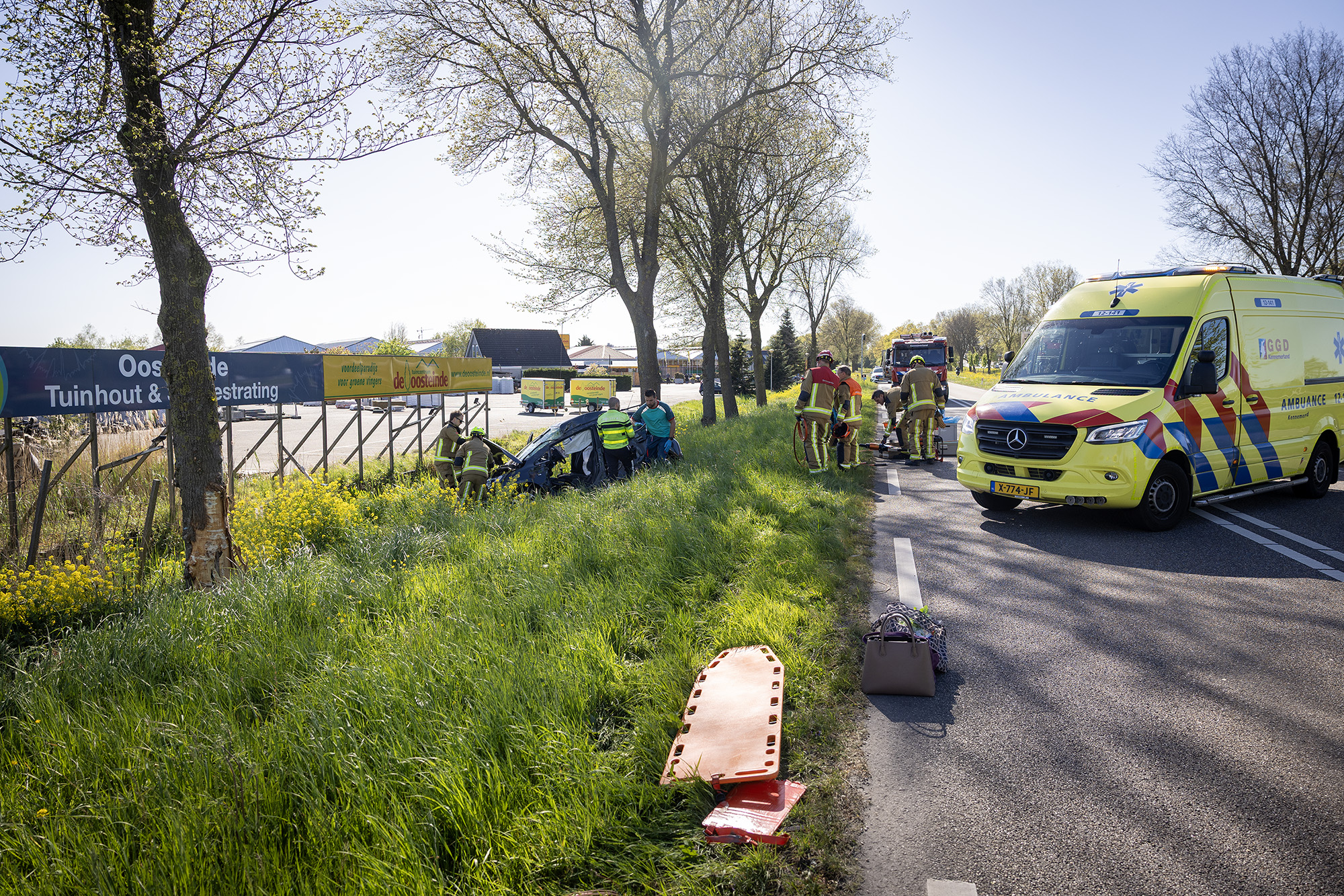 Emergency responders and bystanders attend to a car crash on a rural road as a yellow ambulance sits nearby on the right, with firefighters in gear and a grassy verge in view.