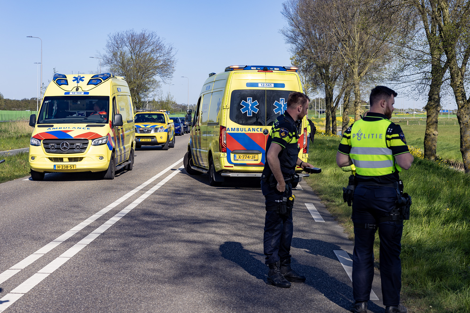 Two yellow ambulances parked on a rural road with police officers standing nearby and a police car behind them, suggesting an emergency scene.