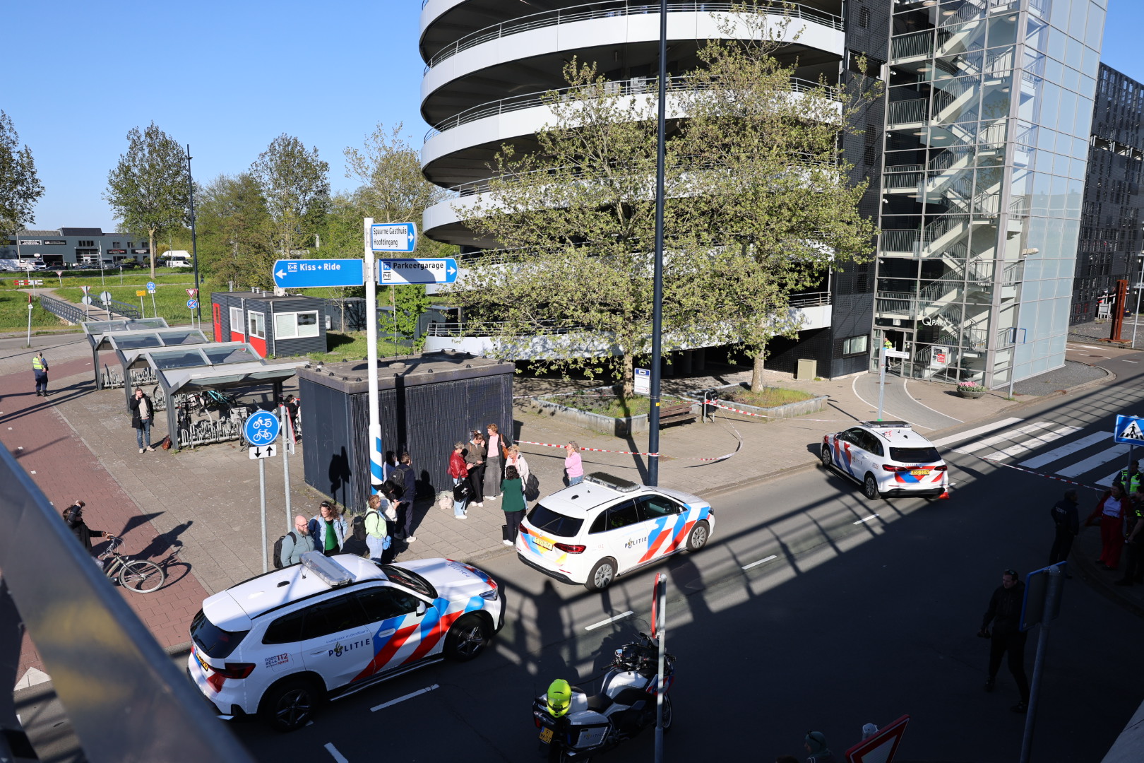 Urban street scene with Dutch police cars patrolling, pedestrians, and cyclists near a modern glass-fronted building far right.