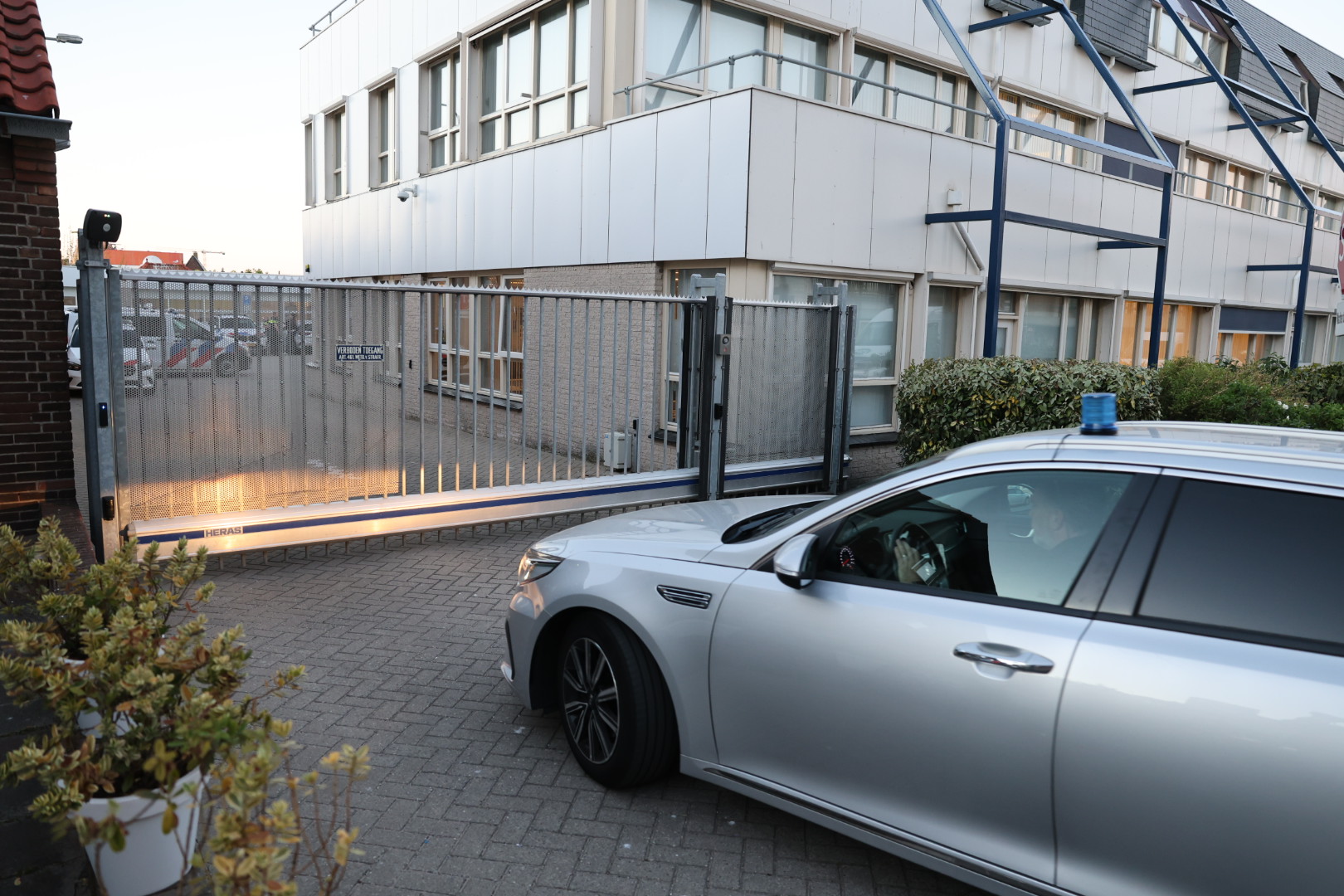 Silver emergency vehicle with blue light bar parking outside a tall metal gate in front of a modern office building.