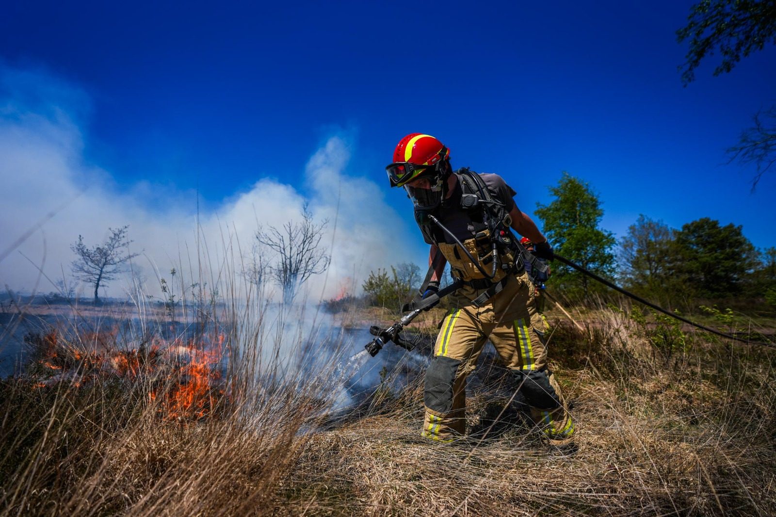 Firefighter in protective gear hoses a grass fire in a dry field under a blue sky.