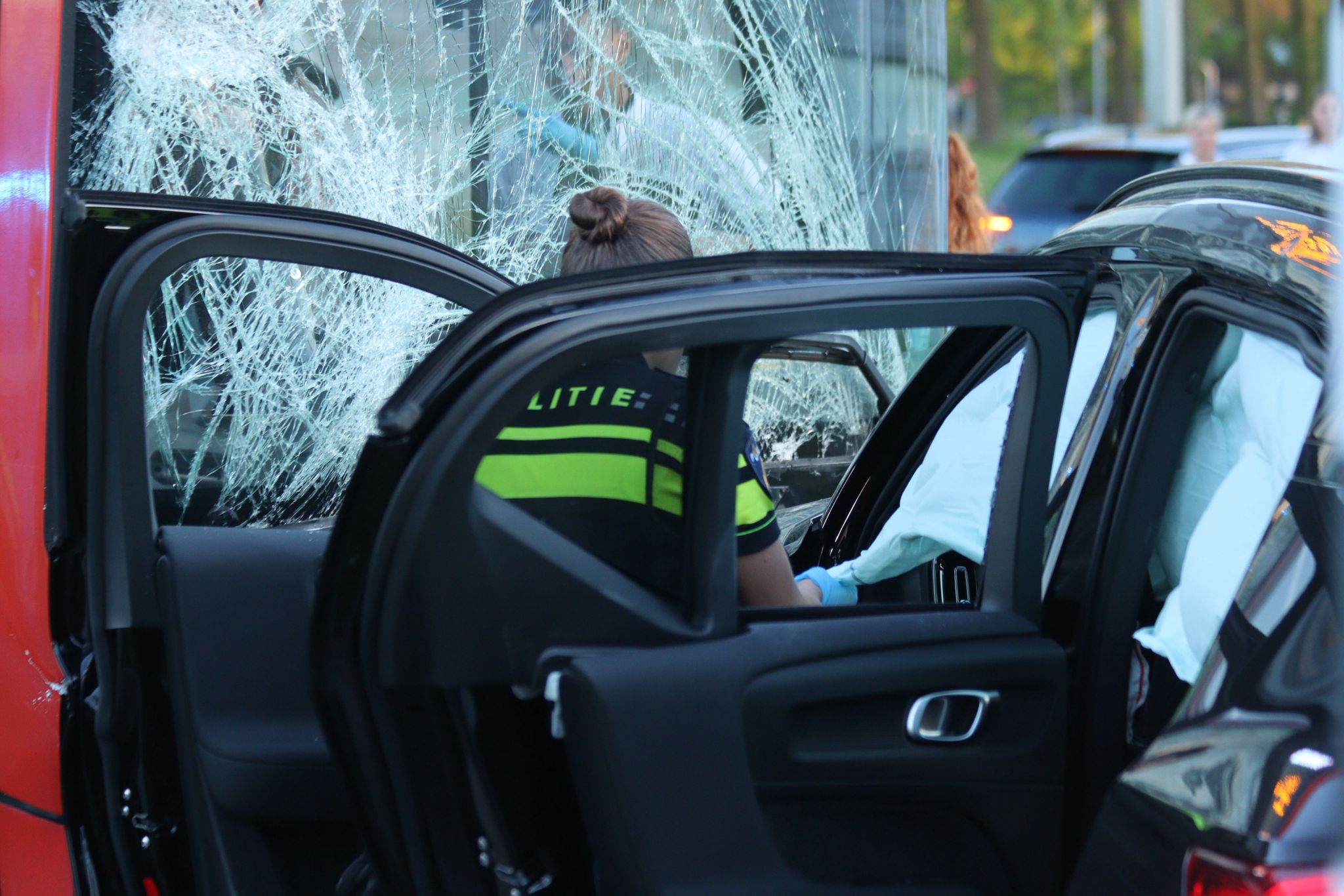 Emergency responder in a neon vest inside a red car after a collision, large shattered windshield visible; bystanders and other vehicles in the background.