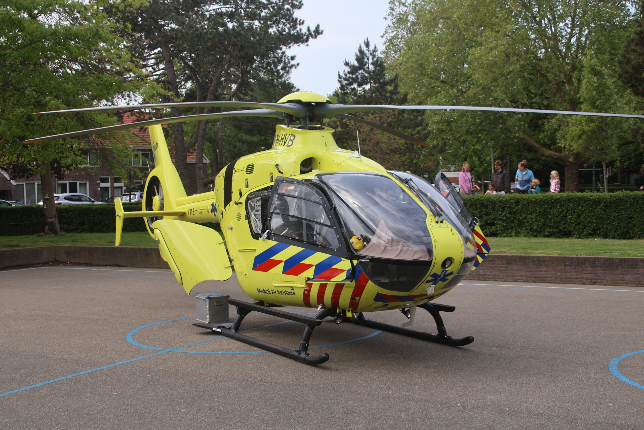 Yellow emergency medical helicopter on a tarmac with onlookers and trees in the background, rotor blades extended.