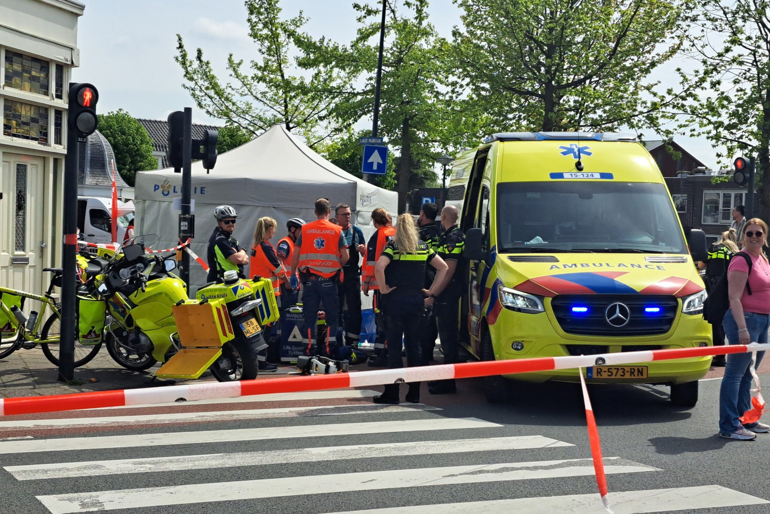 Group of emergency responders in high-visibility vests gather near a bright yellow ambulance on a city street, with a white canopy tent and red-and-white barrier tape in the background.