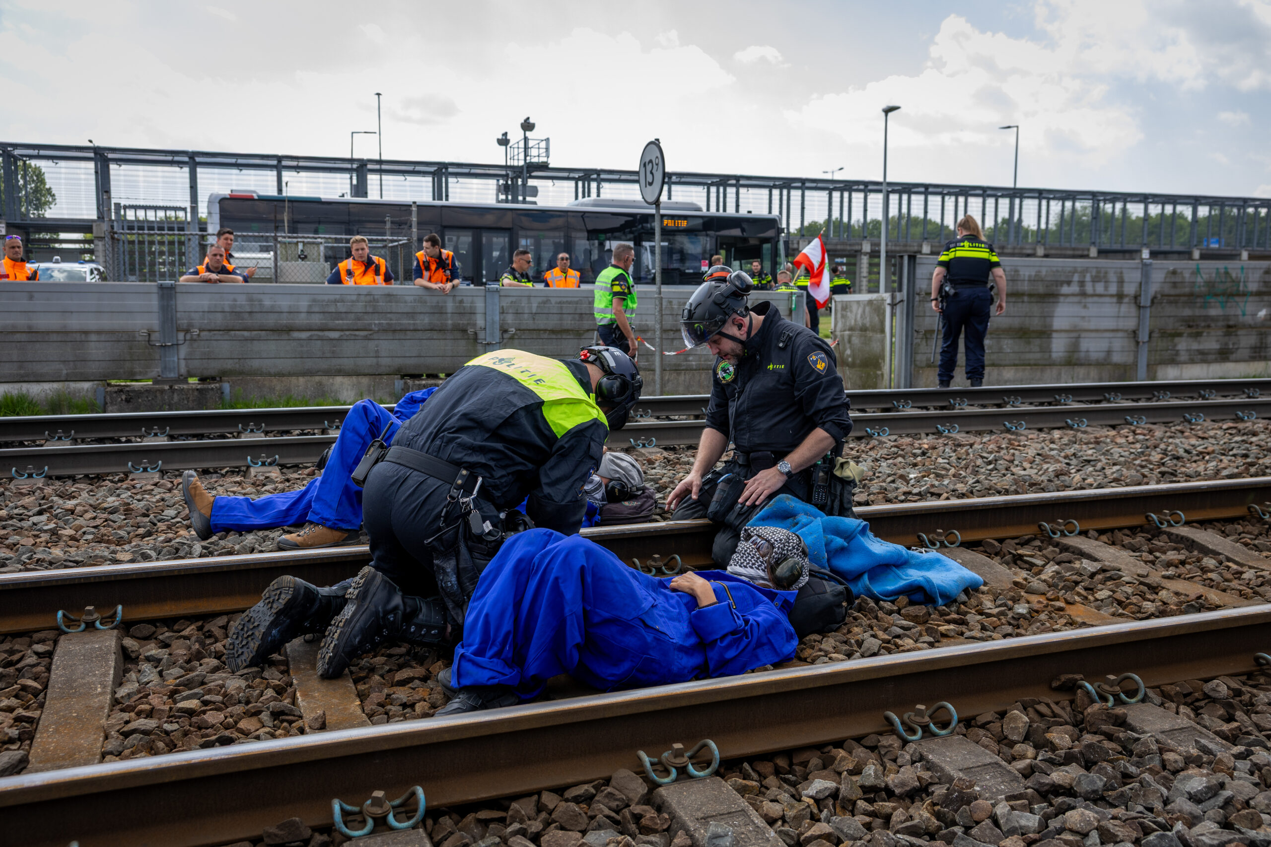 Emergency responders in hi-vis jackets assist a person on the railroad tracks during a drill, with observers behind a fence.