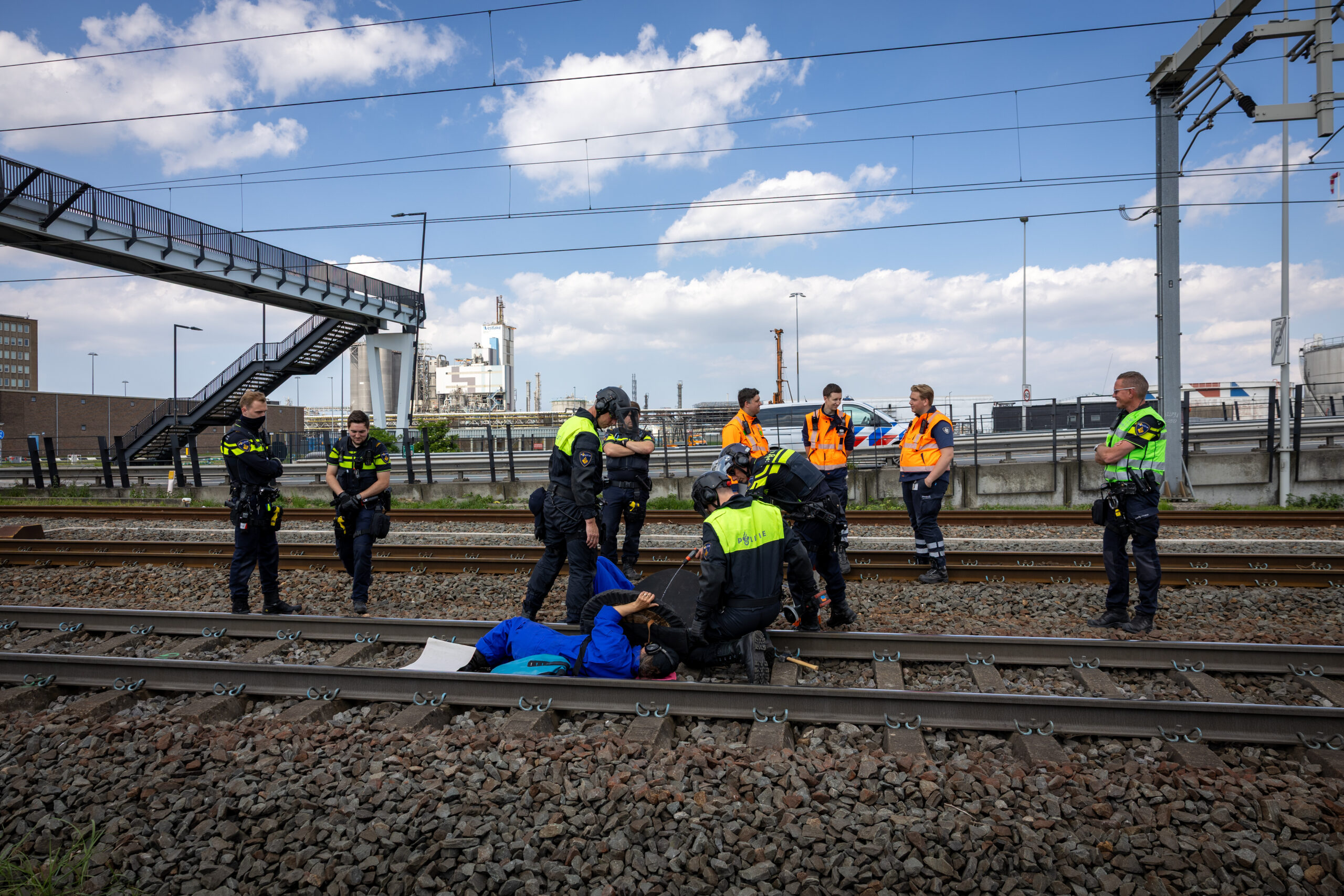 Group of uniformed responders and emergency personnel on and beside railway tracks, some kneeling around equipment, under an overpass with a metal staircase and industrial backdrop.